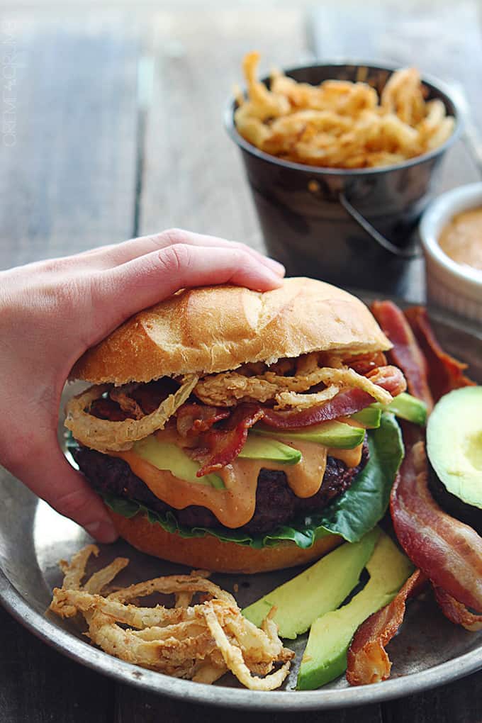 a hand grabbing a western bacon burger with bbq mayo and crispy onion strings from a metal plate.