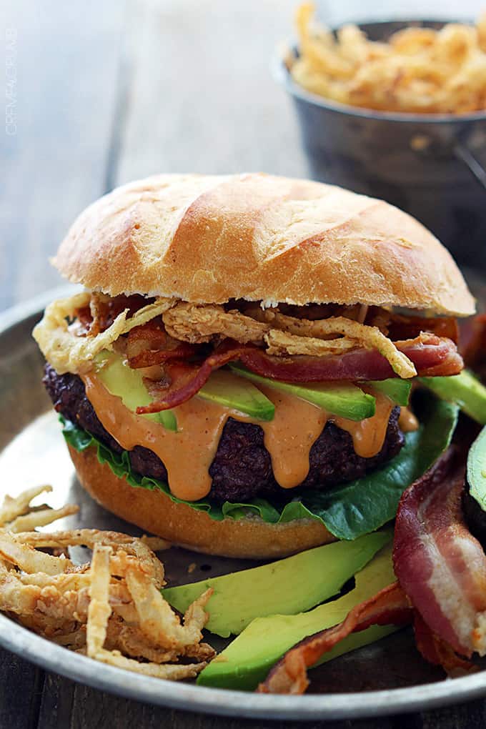 a western bacon burger with bbq mayo and crispy onion strings on a metal plate with more toppings on the side and a bucket of onion strings faded in the background.