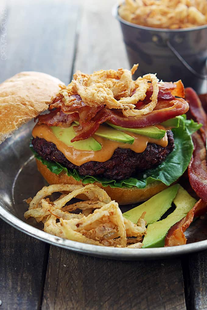 a western bacon burger with bbq mayo and crispy onion strings on a metal plate with a bucket of onion strings faded in the background.