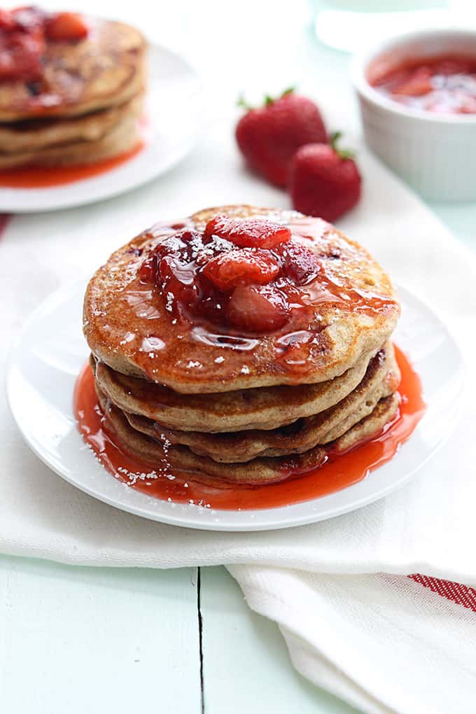 a stack of whole wheat strawberry pancakes on a plate with more pancakes, strawberries, and a bowl of strawberry sauce faded in the background.
