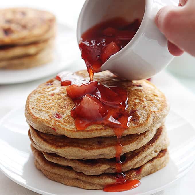 strawberry sauce being poured on top of a stack of whole wheat strawberry pancakes on a a plate with more pancakes faded in the background.