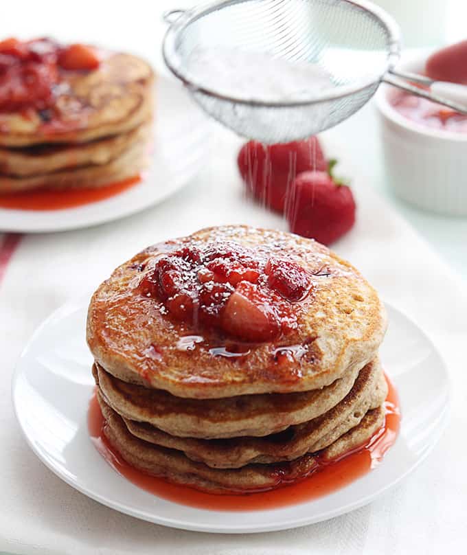 a stack of whole wheat strawberry pancakes on a plate with a hand sifting powder sugar on top.