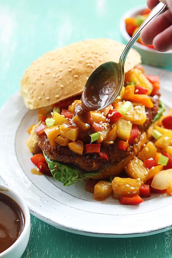 a hand holding a spoon pouring sauce on an an aloha chicken sandwich on a plate.