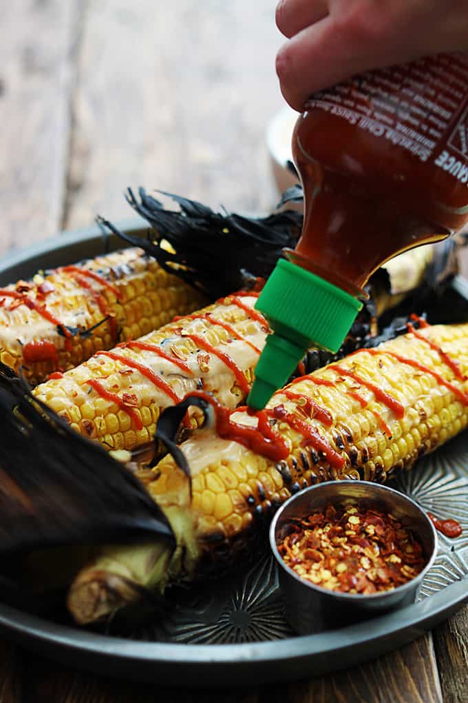 Asian grilled corn on a serving tray with a bowl of seasoning on the side and a hand pouring sriracha sauce on top.