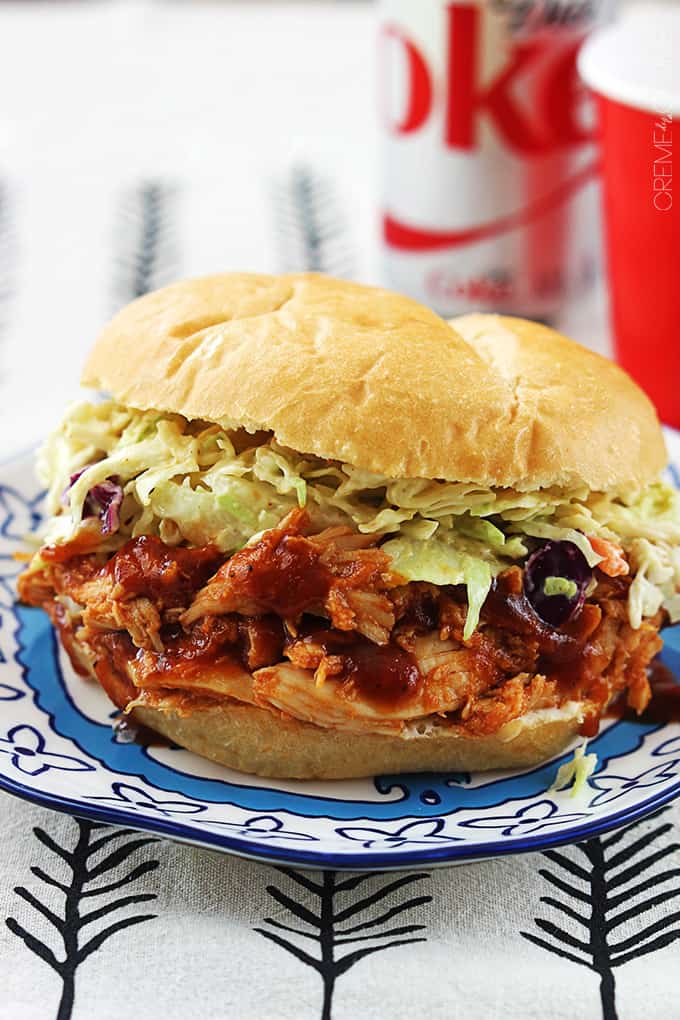 side view of a slow cooker bbq pulled chicken sandwich on a plate with a coke soda can next to a red solo cup faded in the background.