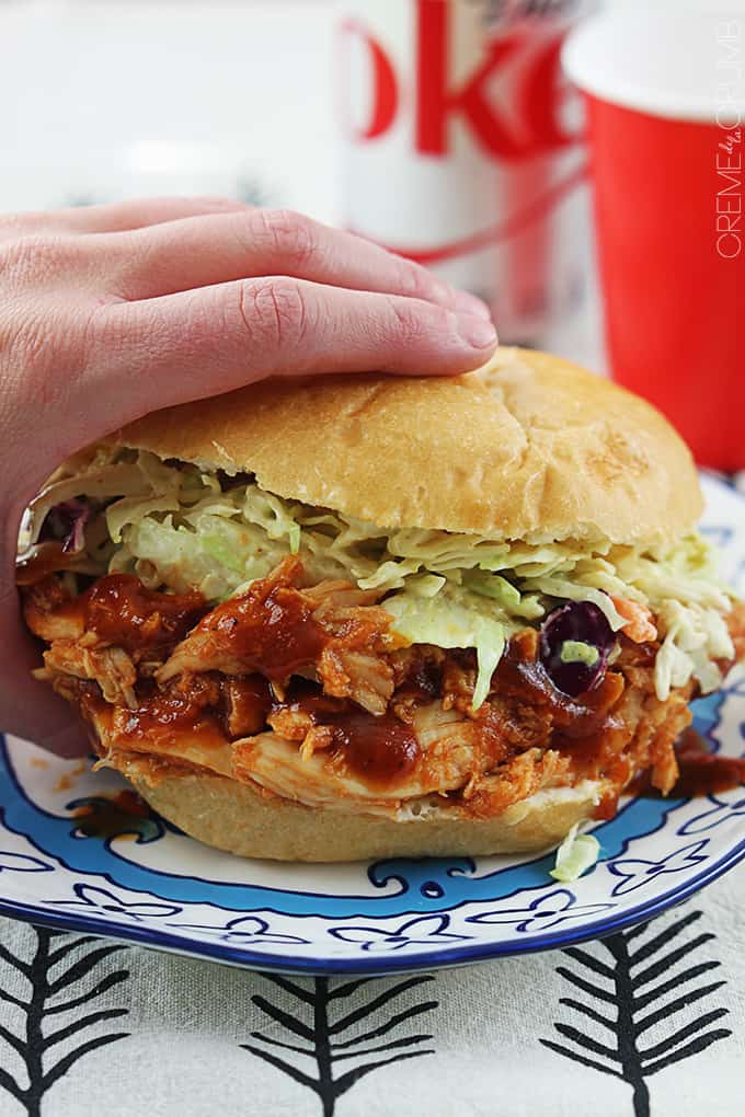 a hand holding on to a slow cooker bbq pulled chicken sandwich on a plate with a coke soda can and a red solo cup faded in the background.