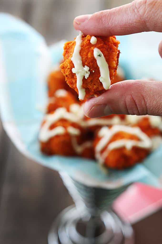 a hand holding a piece of buffalo popcorn chicken with sauce on it with more chicken in a wired cone shaped basket.