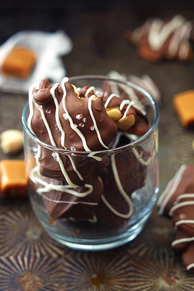 caramel cashew clusters in a small glass with more clusters, caramel pieces, and nuts faded in the background.