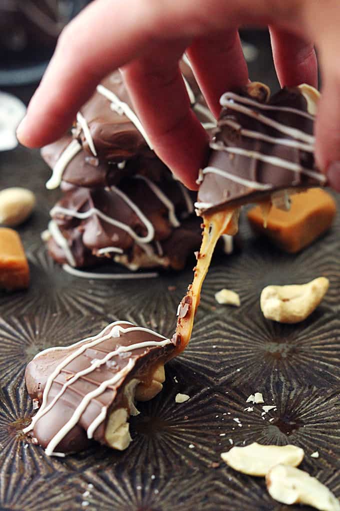a hand pulling a part a caramel cashew cluster with more clusters, caramel pieces, and cashews in the background.