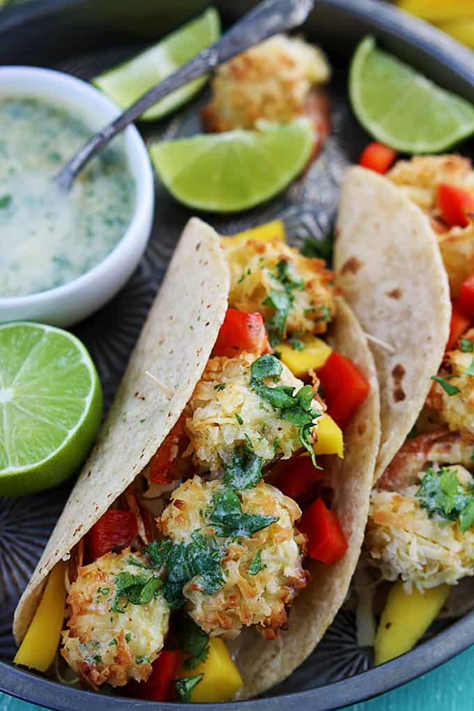 close up of coconut shrimp tacos, slices of lime and sauce on a metal tray.