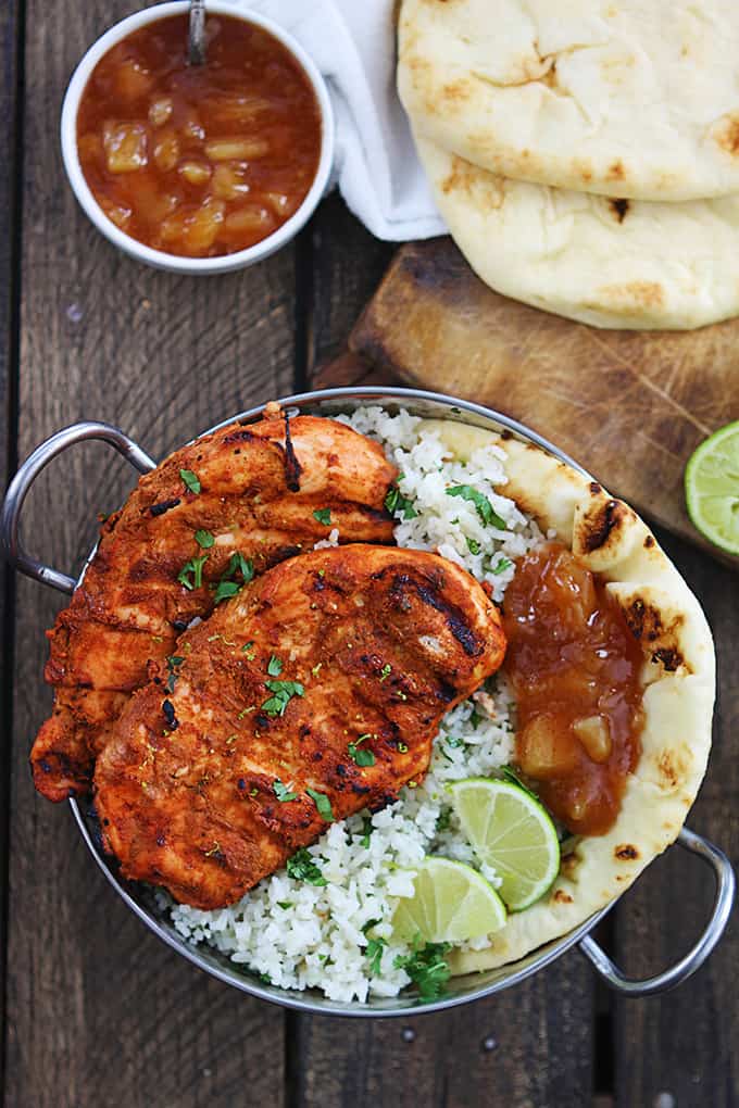 top view of grilled tandoori chicken with rice, pita bread, and limes in a metal tray with more bread and sauce on the side.