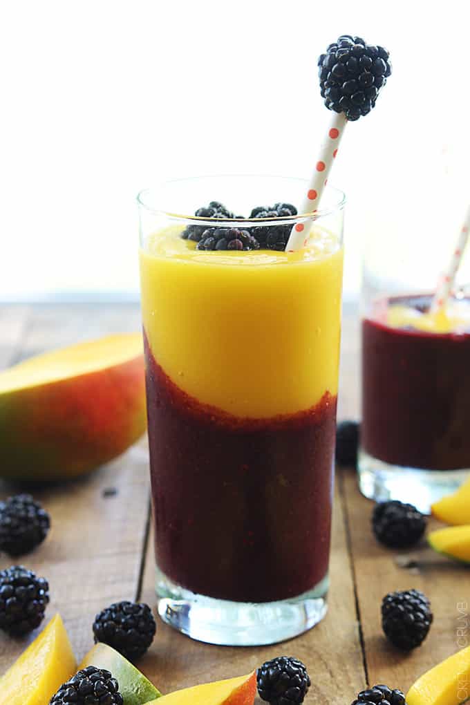 a mango blackberry smoothie in a glass surrounded by blueberries and mangos with another smoothie in the background.