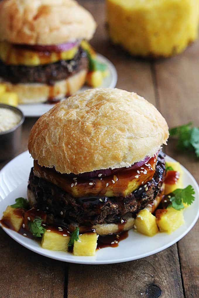 a black bean teriyaki burger on a plate with another burger on a plate and a pineapple faded in the background.