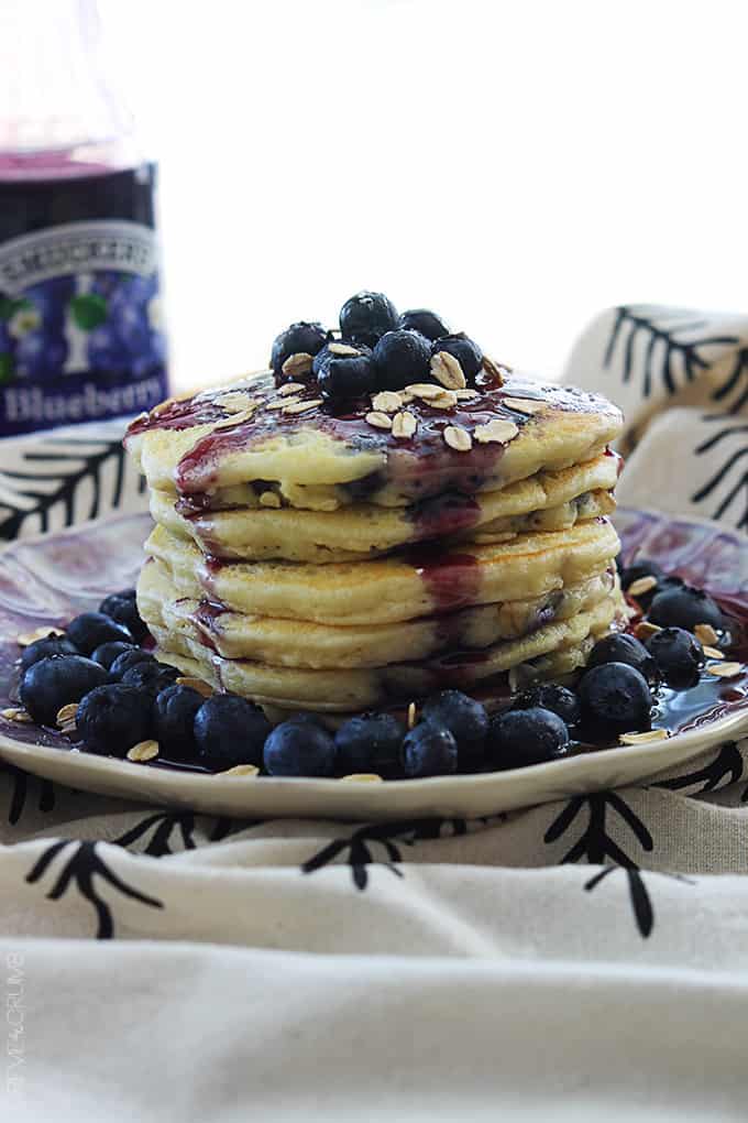 a stack of blueberry oatmeal pancakes with oats, blueberries, and blueberry syrup on top on a plate.