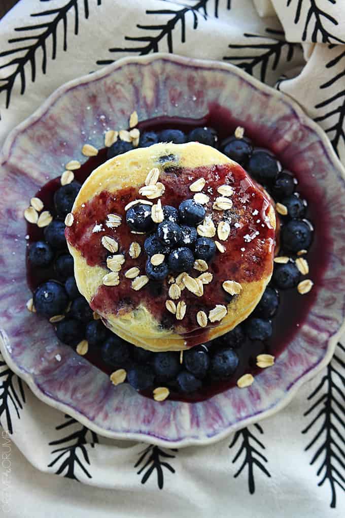 top view of a stack of blueberry oatmeal pancakes topped with oats, blueberries, and blueberry syrup on a plate.