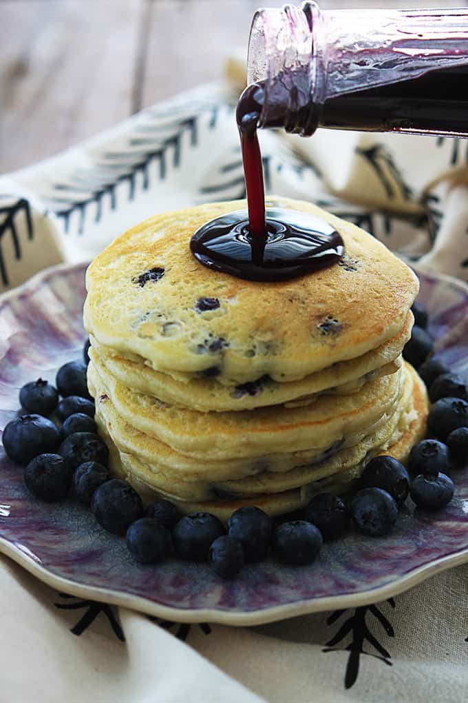 a stack of blueberry oatmeal pancakes surrounded by blueberries on a plate with blueberry syrup being poured on top.