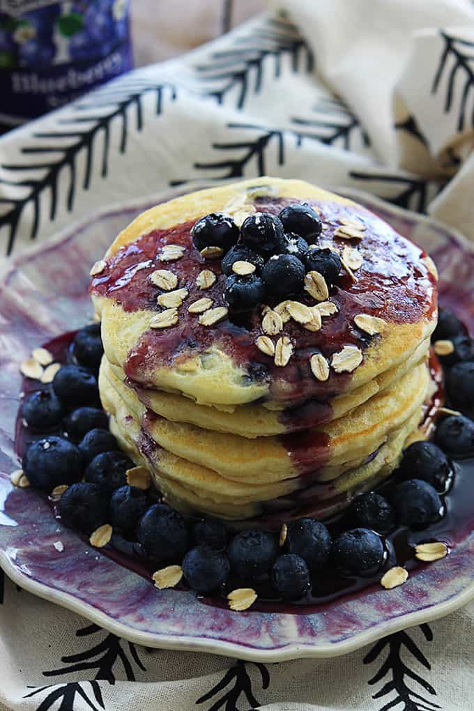 a stack of blueberry oatmeal pancakes topped with blueberries, oats and blueberry syrup on a plate.