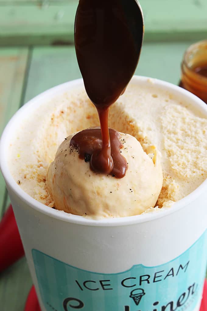 a spoon pouring sauce on top of a scoop of butterscotch ice cream in a container of ice cream.