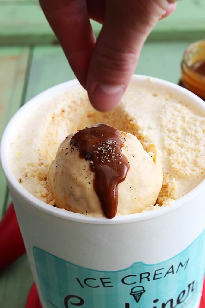 a hand sprinkling sea salt on sauce on a scoop of butterscotch ice cream in a container of ice cream.
