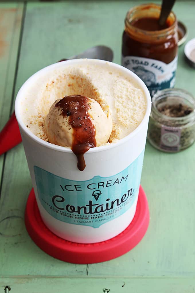 a container of butterscotch ice cream with sauce on a scoop of ice cream and jars of toppings and an ice cream scooper in the background.