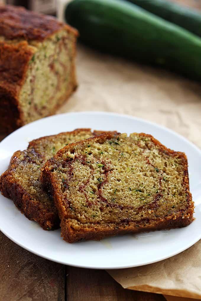 slices of cinnamon swirl zucchini bread on a plate with a loaf of bread and zucchinis in the background.