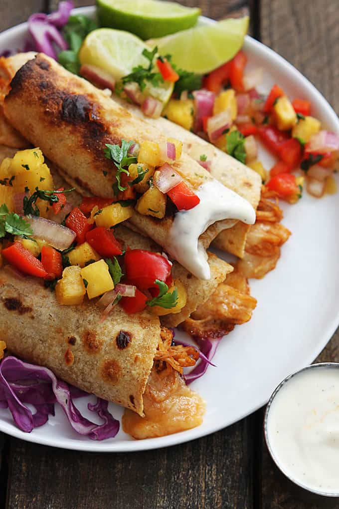 top view of slow cooker Hawaiian bbq chicken taquitos topped with pineapple salsa on a plate with the top taquito dipped in ranch with a bite taken out.