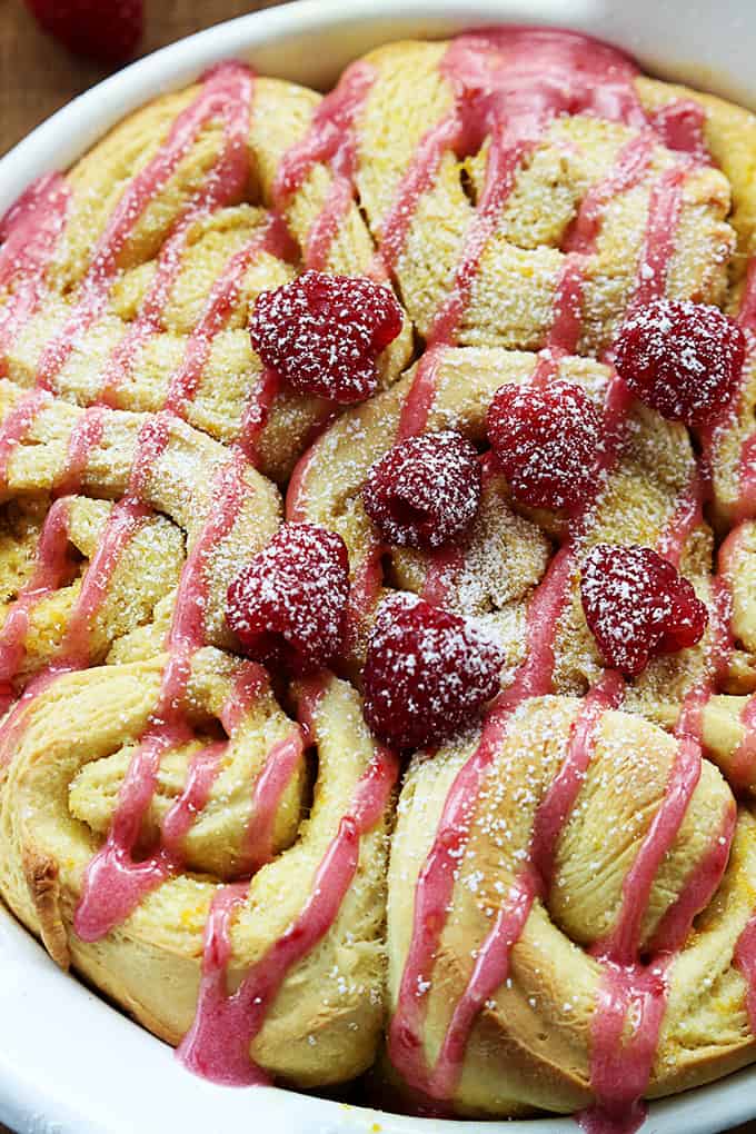 close up top view of orange raspberry sweet rolls in a serving tray topped with icing and raspberries.