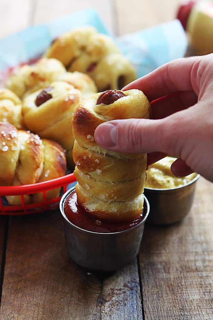 a hand dipping a pretzel dog in ketchup with more pretzel dogs in a basket.