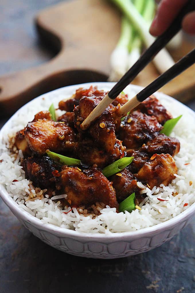 a hand holding chopsticks taking some general tso's chicken from a bowl of chicken and rice.