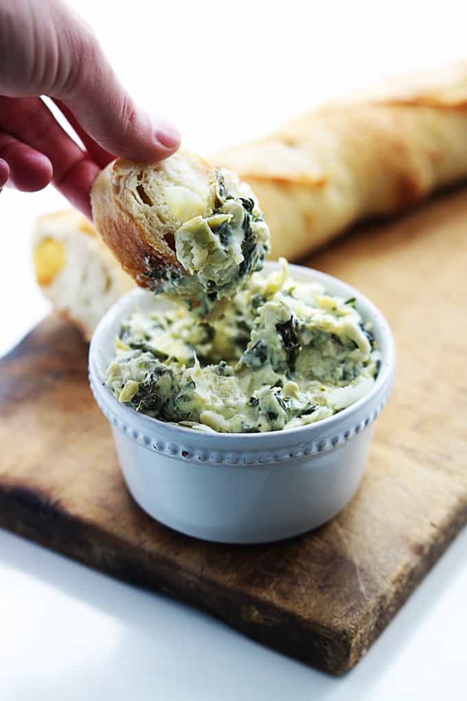 a hand lifting a piece of bread with slow cooker spinach artichoke dip from the bowl of dip.