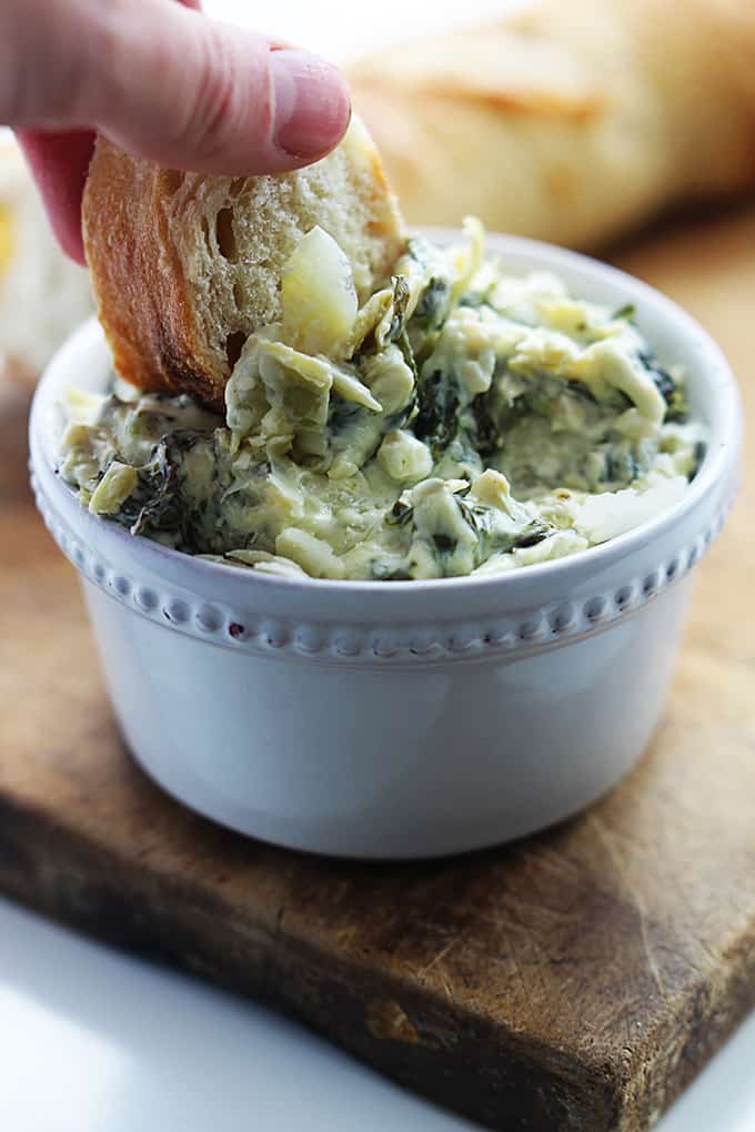 a hand dipping a piece of bread in a bowl of slow cooker spinach artichoke dip.
