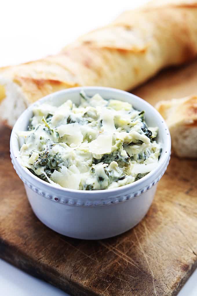 a bowl of slow cooker spinach artichoke dip with a load of bread in the background all on top of a wooden cutting board.