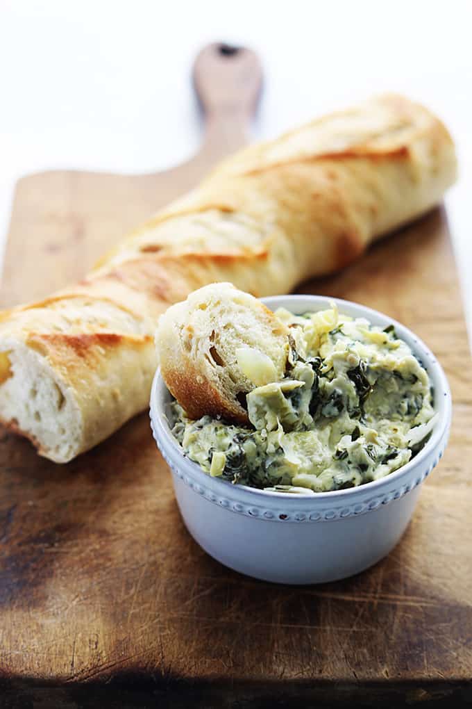 slow cooker spinach artichoke dip in a bowl with a slice of bread dipped in it with the rest of the loaf behind it all on a wooden cutting board.