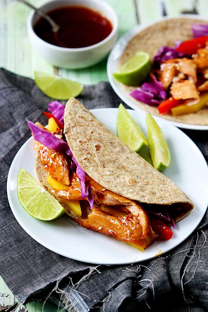 a Thai salmon taco on a plate with slices of lime and sauce and a another taco in the background.
