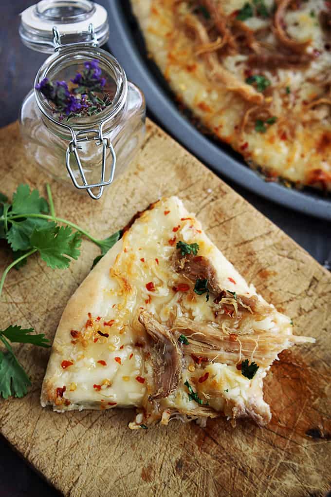 a slice of Thai sweet chili pork pizza with a bite missing on a wooden cutting board with a jar of spices next to it and the rest of the pizza on the side.