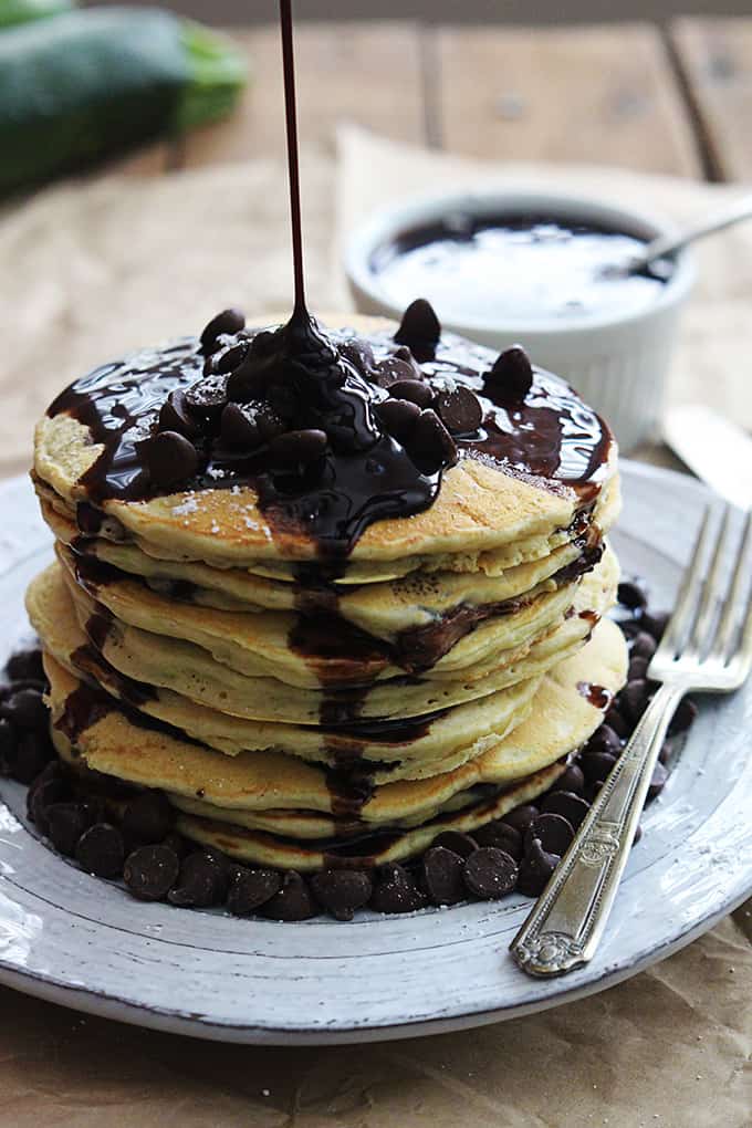 a stack of zucchini chocolate chip pancakes with a fork on the side and chocolate syrup being poured on the top.