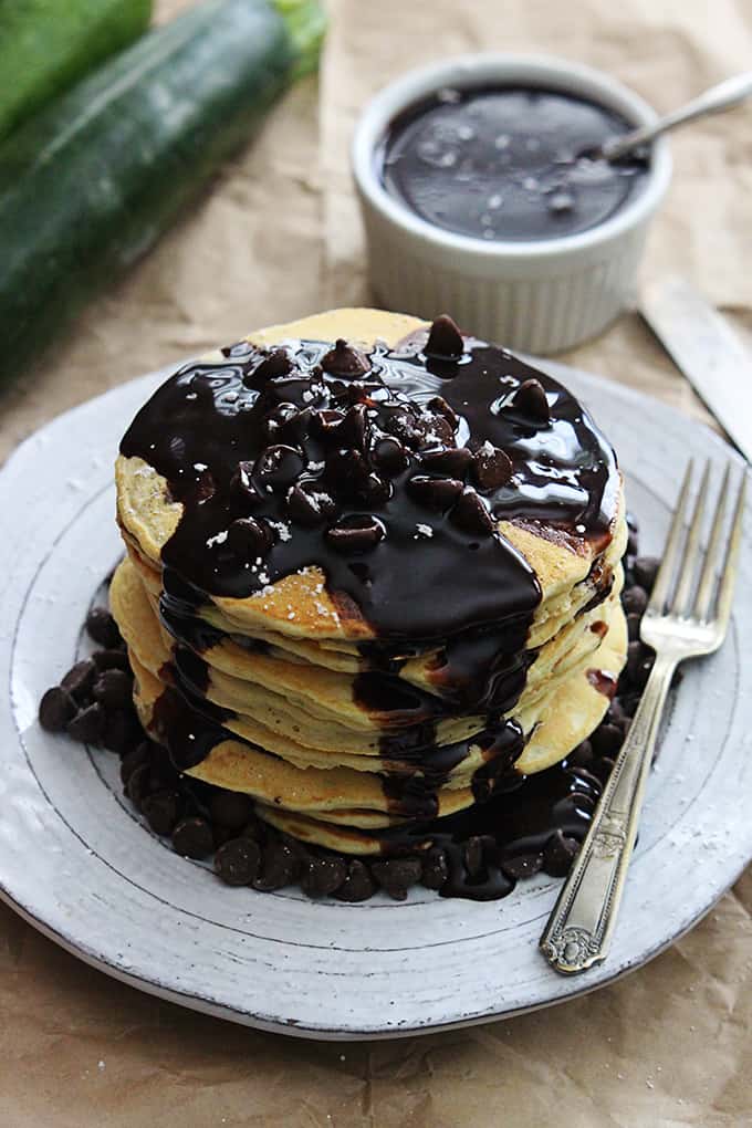a stack of zucchini chocolate chip pancakes topped with chocolate chips and chocolate syrup on a plate with a fork on the side and more syrup and zucchini in the background.