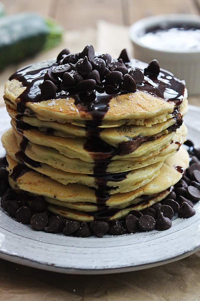 a stack of zucchini chocolate chip pancakes on a plate with chocolate chips and chocolate syrup.