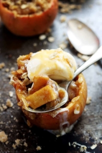 an apple crisp stuffed apple topped with vanilla ice cream with a spoon with another spoon and apple in the background.