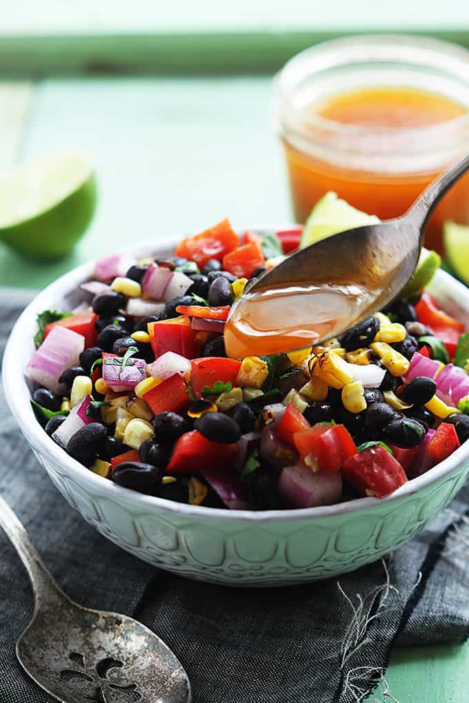 a spoon pouring honey on top of honey chipotle black bean salad with a slice of lime in a bowl.