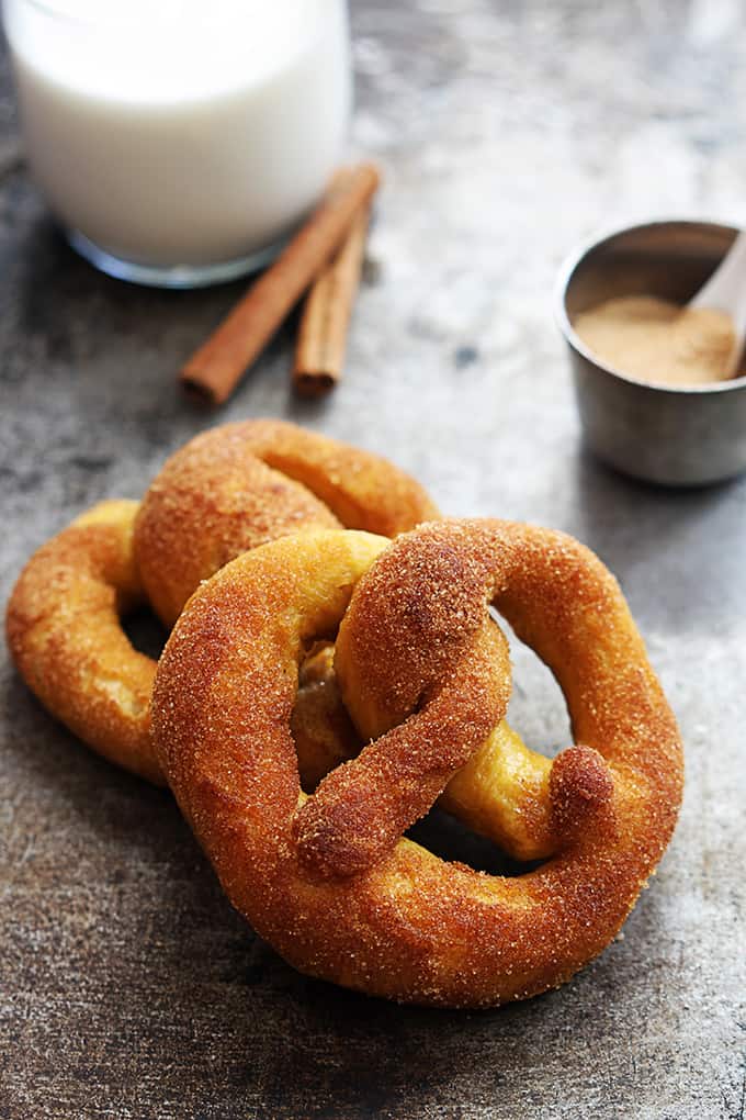 two cinnamon sugar pumpkin soft pretzels leaning on top of each other with a glass of milk, cinnamon sticks and a small bowl of cinnamon sugar in the background.