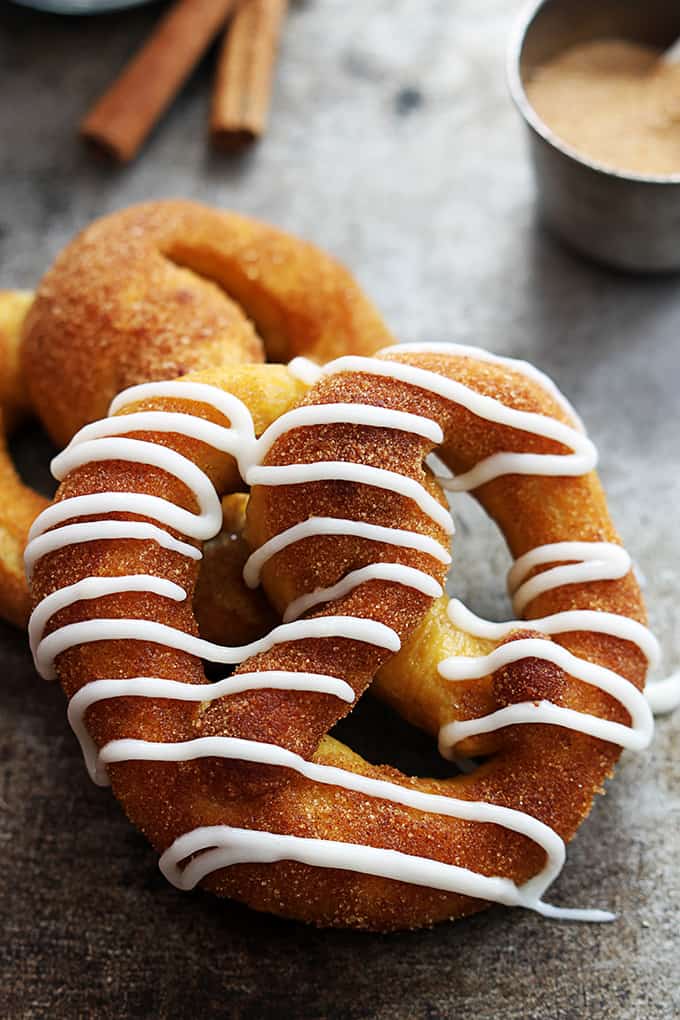 cinnamon sugar pumpkin soft pretzels leaning on top of each other with the top pretzel being iced with vanilla icing with cinnamon sticks and a small bowl of cinnamon sugar in the background.