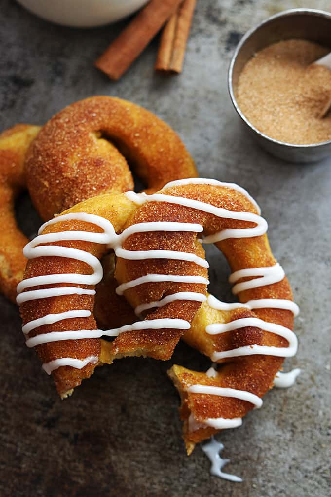 top view of two cinnamon sugar pumpkin soft pretzels on top of each other with the top pretzel having vanilla icing on top and missing a bite with a glass of milk, cinnamon sticks, and a small bowl of cinnamon sugar on the side.