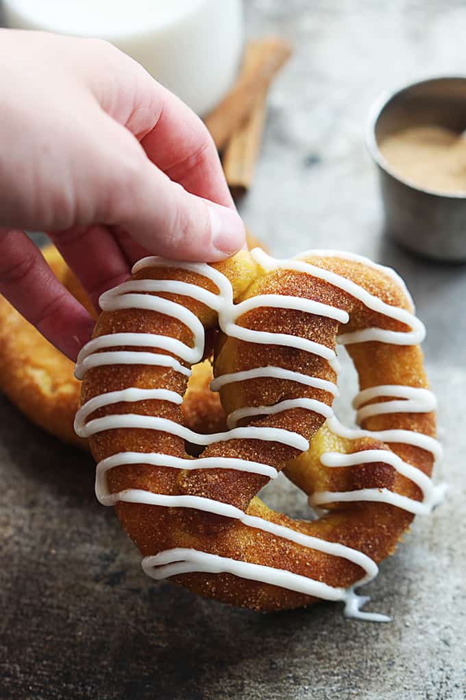 a hand picking up a cinnamon sugar pumpkin soft pretzels iced with vanilla icing with another pretzel, cinnamon sticks, a small bowl of cinnamon sugar and milk faded in the background.