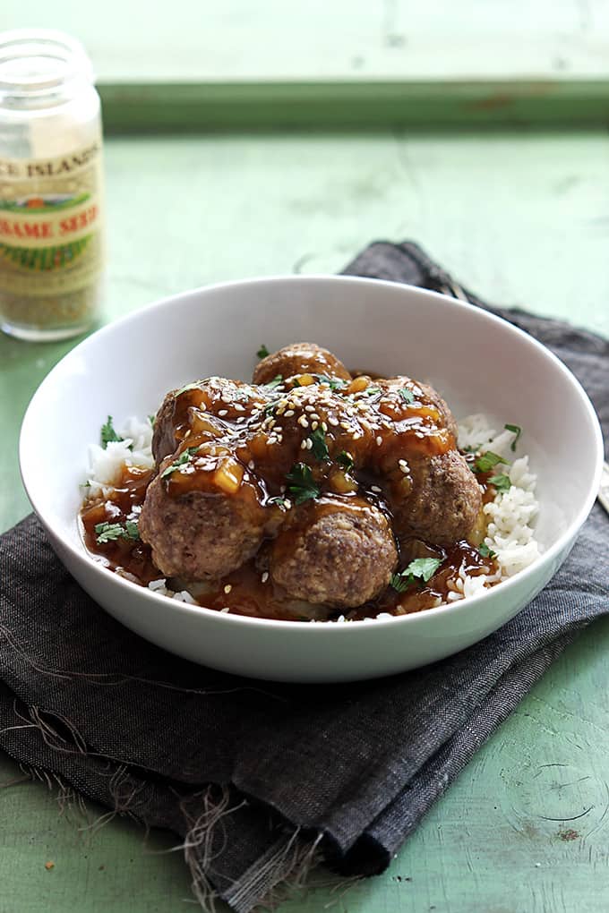 Hawaiian meatballs on rice in a bowl on to pot a cloth napkin with a jar of sesame seeds on the side.