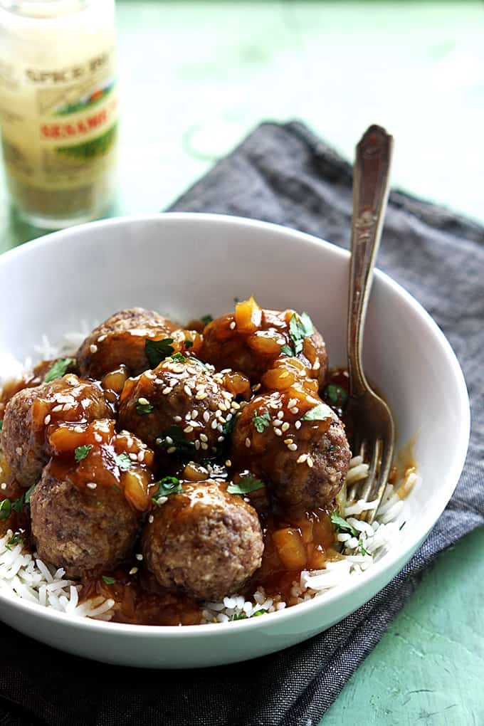 Hawaiian meatballs on rice with a fork in a bowl on a cloth napkin with a jar of sesame seeds on the side.