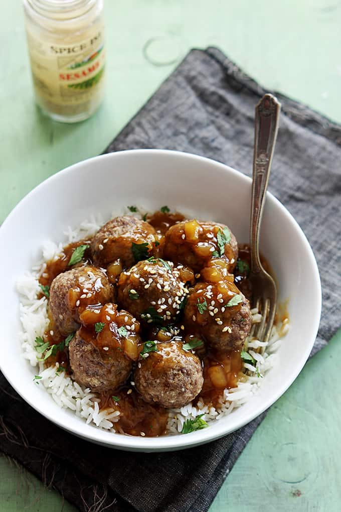 top view of Hawaiian meatballs on rice with a fork in a bowl on a cloth napkin with a jar of sesame seeds on the side.