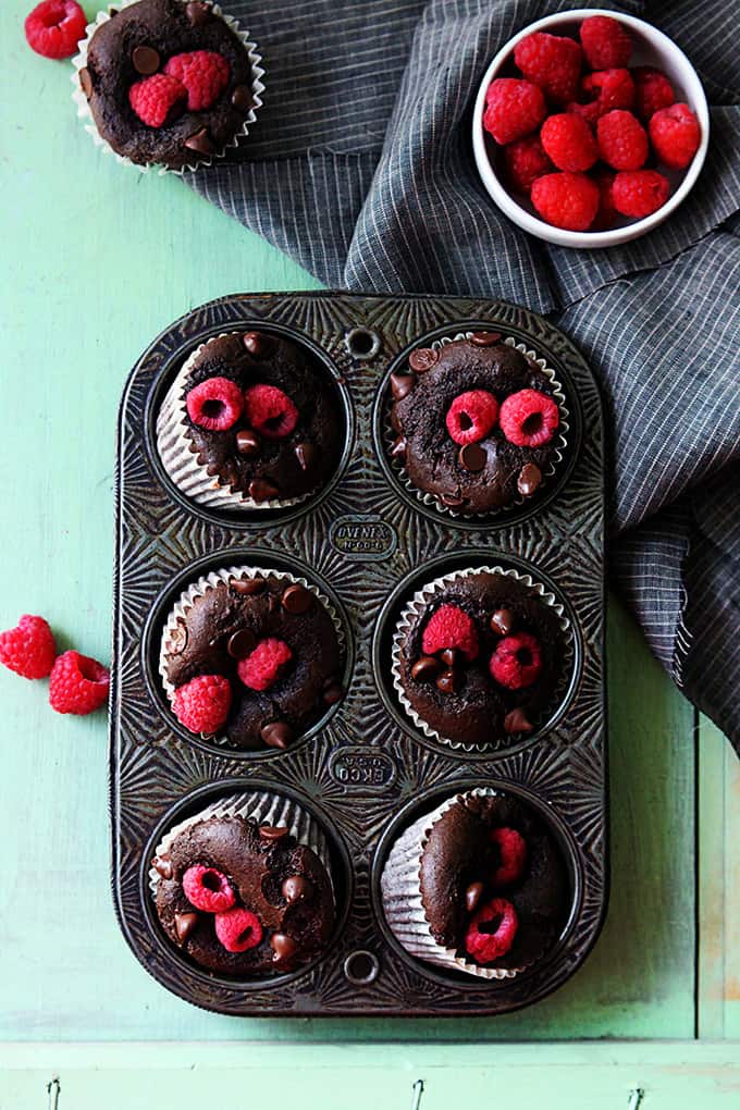 top view of raspberry chocolate muffins in a muffin pan with a bowl of raspberries, another muffin, and more raspberries on the side.