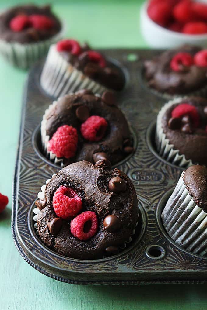 close up of raspberry chocolate muffins in a muffin pan with another muffin and bowl of raspberries faded in the background.