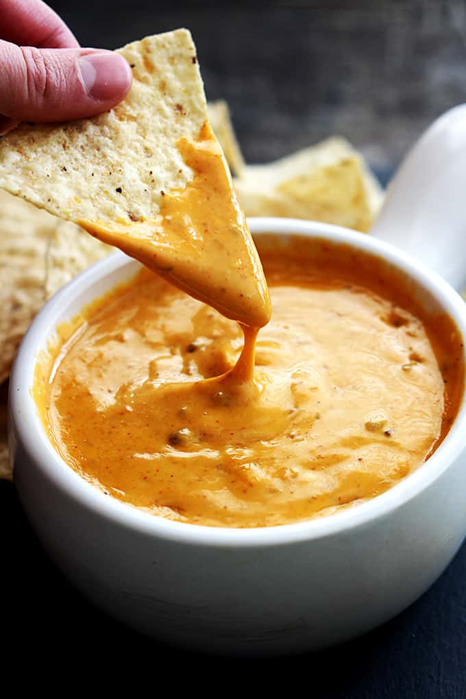 a hand holding a chip just dipped in Chili's queso (slow cooker version) with the bowl of queso underneath and more chips in the background.